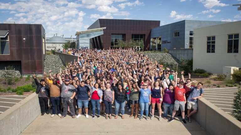 grande grupo de estudantes reunidos ao ar livre no campus, sorrindo e levantando os braços em celebração, com prédios modernos ao fundo.